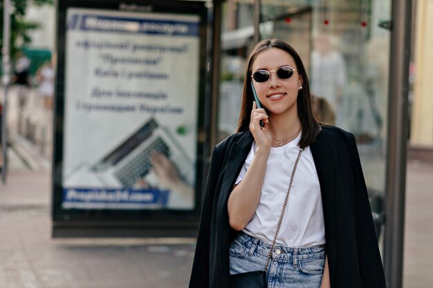어두운 재킷 흰색 셔츠와 안경을 쓴 세련된 독립 여성이 행복한 미소로 시내 거리에서 전화 통화를 하고 있습니다
