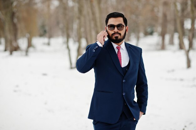 スーツとサングラスのスタイリッシュなインドのひげのビジネスマンは、携帯電話で話す冬の日の屋外でポーズをとった