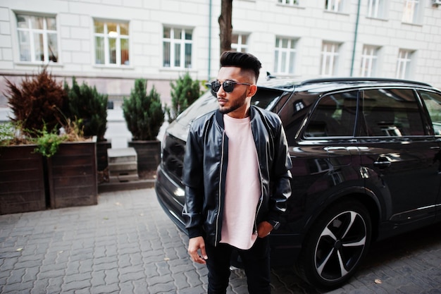 Elegante uomo con barba indiana in giacca di pelle nera e occhiali da sole contro il modello indiano dell'auto suv d'affari posato all'aperto nelle strade della città