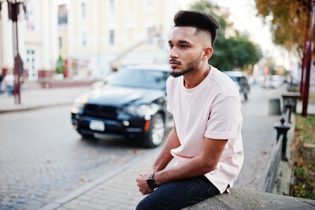 Stylish indian beard man at pink tshirt sitting against black suv car India model posed outdoor at streets of city