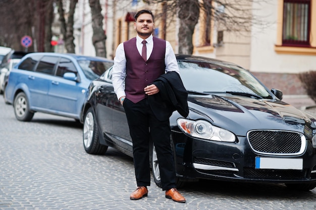 Stylish indian businessman in formal wear vest suit standing against black business car on street of city
