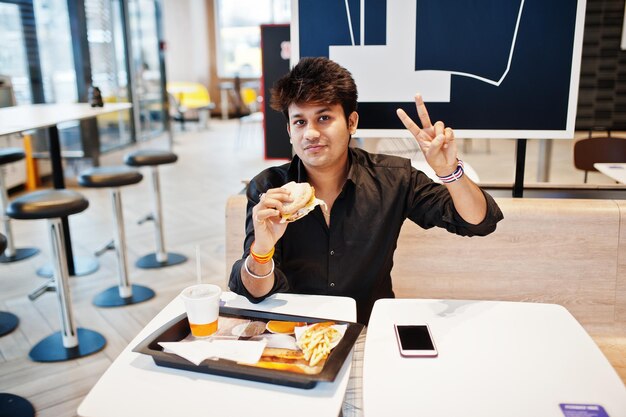 Stylish indian man sitting at fast food cafe and eating hamburger and gesture peace sign hand