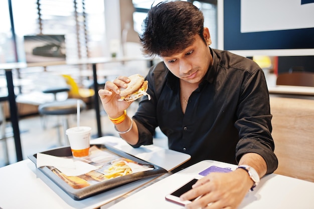 Stylish indian man sitting at fast food cafe and eating hamburger read morning news on mobile phone and have surprised eyes