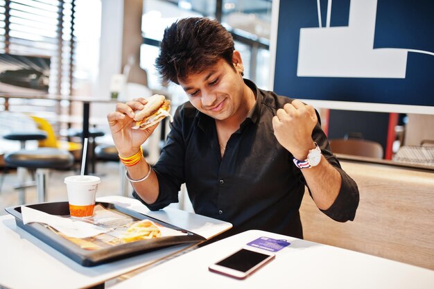 Stylish indian man sitting at fast food cafe and eating hamburger read morning news on mobile phone He is happy and show like him win something