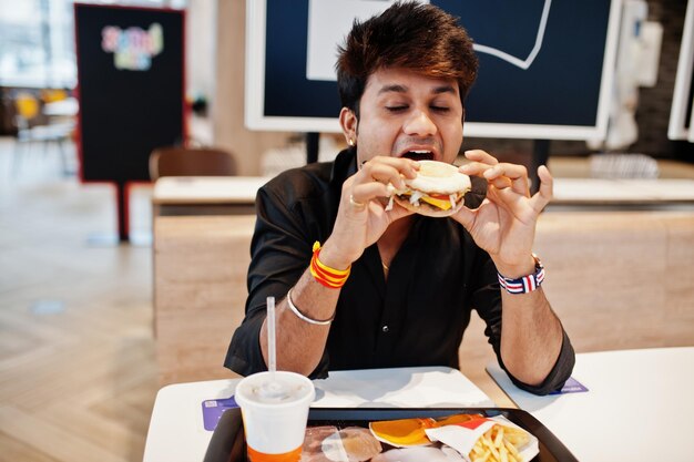 Stylish indian man sitting at fast food cafe and eating hamburger