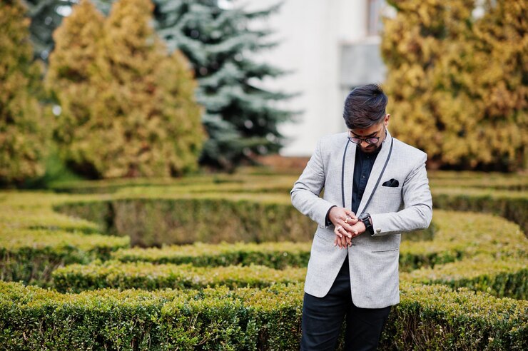 Free Photo stylish indian man with bindi on forehead and glasses wear on suit posed outdoor against green bushes at park and looking at his watches