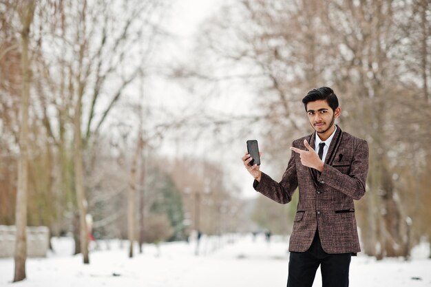 갈색 양복과 안경을 쓴 세련된 인도 학생 남자는 겨울날 야외에서 휴대전화를 들고 포즈를 취했습니다.