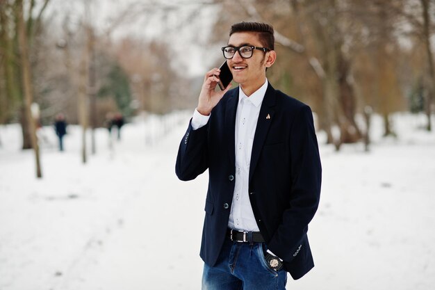 양복과 안경을 쓴 세련된 인도 학생 남자는 겨울날 야외에서 포즈를 취하고 휴대전화로 이야기합니다.