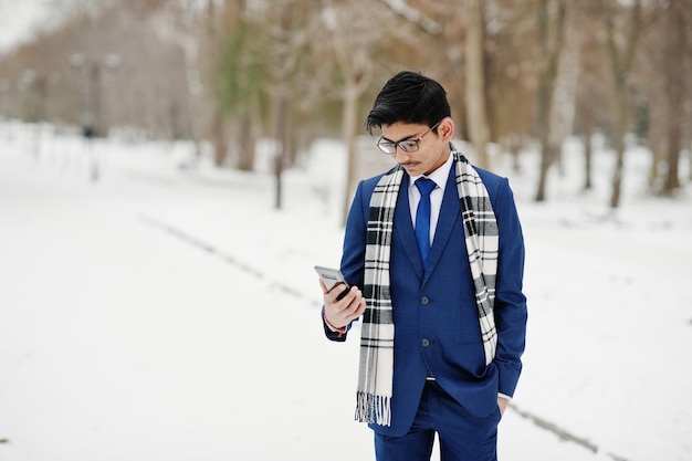 携帯電話を見て冬の日の屋外でポーズをとったスーツのメガネとスカーフのスタイリッシュなインドの学生男性