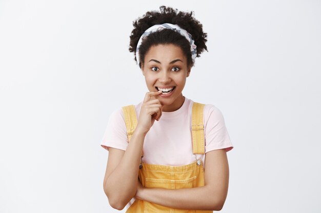 stylish intrigued and cute african woman with trendy headband in yellow dungarees, biting finger while curiously listening to interesting story, smiling broadly and gazing