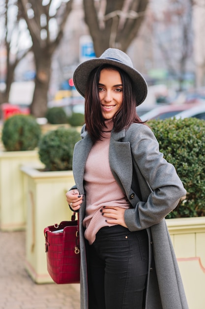 Stylish joyful young woman in long grey coat, hat walking on street in city on park. Luxury clothes, fashionable model, smiling, cheerful mood, elegant outlook.
