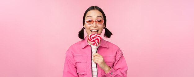 사탕을 먹고 분홍색 배경에 선글라스를 끼고 웃고 있는 사탕을 핥는 세련된 한국 소녀