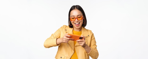 Free photo stylish korean girl in sunglasses playing mobile video game laughing and smiling while using smartphone standing over white background