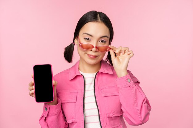 Stylish korean girl young woman in sunglasses showing smartphone screen mobile phone app interface or website standing over pink background