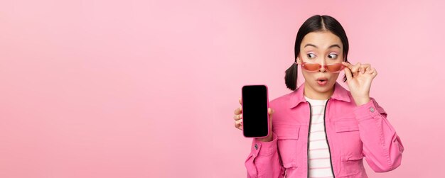 Stylish korean girl young woman in sunglasses showing smartphone screen mobile phone app interface or website standing over pink background