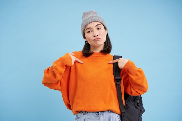 Free photo stylish korean woman points at herself stands in warm hat and holds backpak on shoulder blue backgro