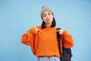 Free photo stylish korean woman points at herself stands in warm hat and holds backpak on shoulder blue backgro