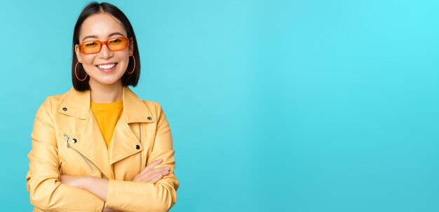 Stylish korean woman in sunglasses and yellow jacket cross arms smiling and looking confident at cam