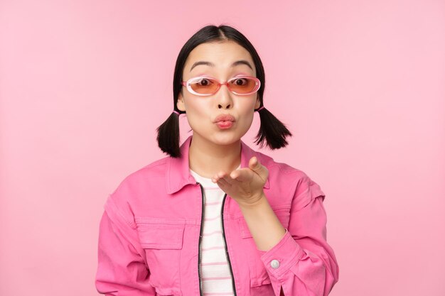 선글라스를 쓴 세련된 케이팝 소녀 아시아 여성이 분홍색 스튜디오 배경 위에 서 있는 카메라 주름 입술 mwah 제스처에 공기 키스를 부는