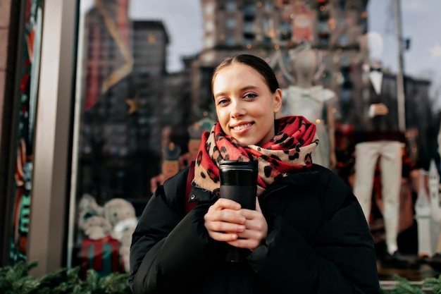 Free photo stylish lady in great mood standing on window with black wooden frame and holding coffee girl dressed in warm jacket scarf