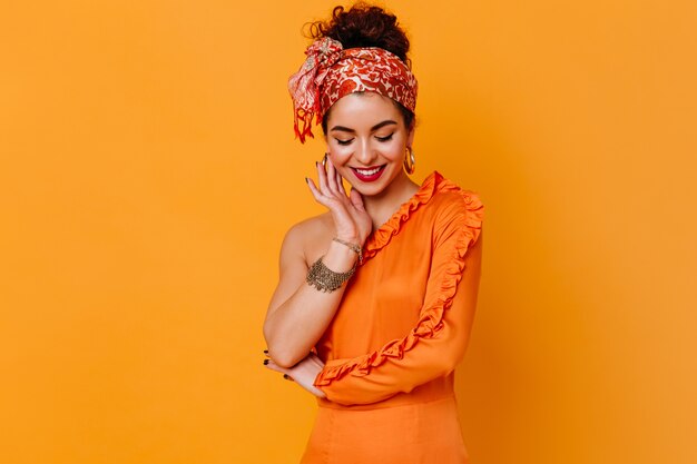 Stylish lady in orange dress and bright bandage on her head with shy smile looks down on orange space.