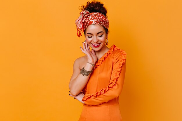 Стильная дама в оранжевом платье и яркой повязке на голове с застенчивой улыбкой смотрит на оранжевое пространство.