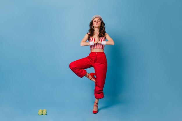 Stylish lady in red pants and striped cropped top standing in tree pose on blue wall with green dumbbells