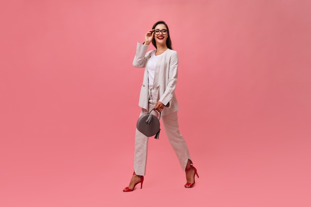 Stylish lady in suit holds handbag and walks on pink background.  Business woman with dark hair with bright red lips and stylish heels smiling.
