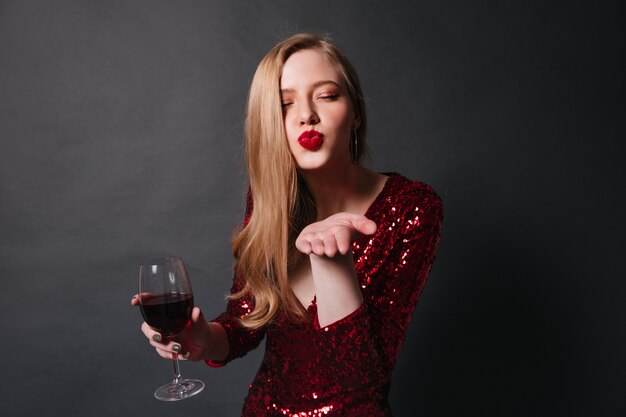エアキスを送信するワイングラスを持つスタイリッシュな女性。パーティーでワインを飲む赤いドレスを着たブロンドの女の子のスタジオショット。