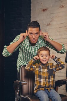 Стильный маленький ребенок сидит на стуле в парикмахерской со своим молодым отцом на заднем плане, они дурачатся и веселятся