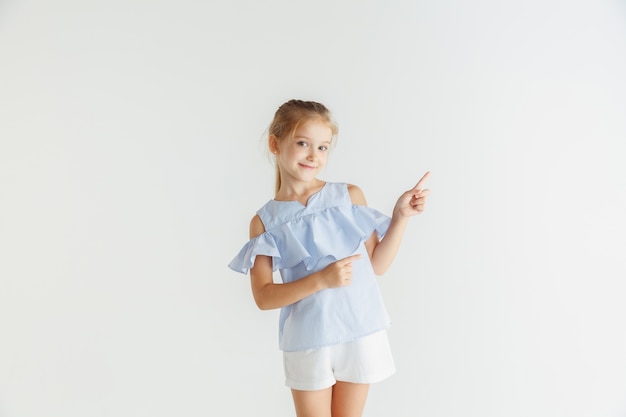 Stylish little smiling girl posing in casual clothes isolated on white studio background. Caucasian blonde female model. Human emotions, facial expression, childhood. Pointing on empty space bar.