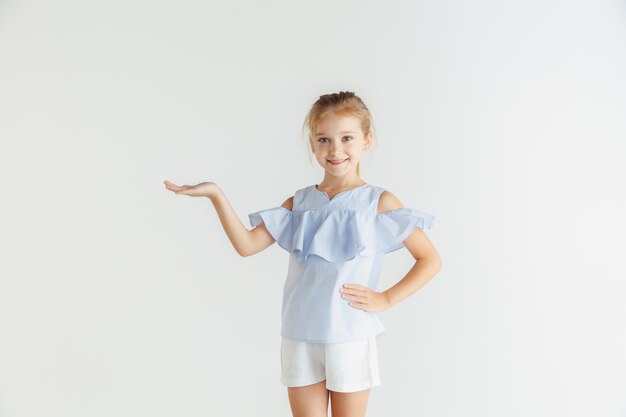 Stylish little smiling girl posing in casual clothes isolated on white studio background. Caucasian blonde female model. Human emotions, facial expression, childhood. Showing an empty space.