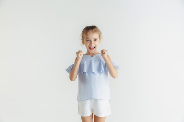 Stylish little smiling girl posing in casual clothes isolated on white wall. Caucasian blonde female model. Human emotions, facial expression, childhood. Winning, celebrating, smiling.