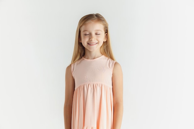 Stylish little smiling girl posing in dress isolated on white studio background. Caucasian blonde female model. Human emotions, facial expression, childhood. Looks calm, smiling with eyes closed.