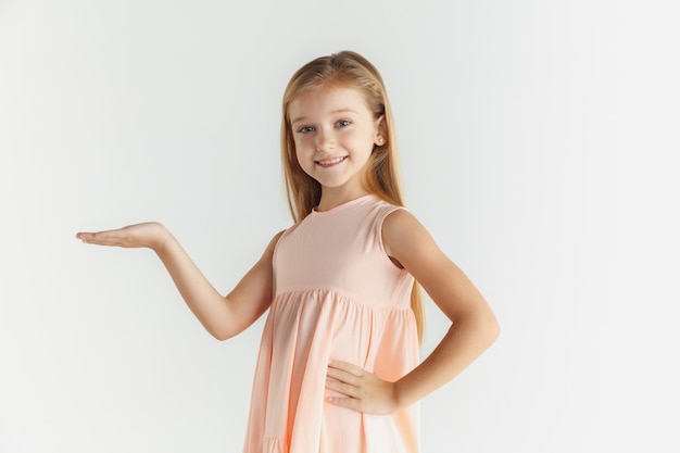 Stylish little smiling girl posing in dress isolated on white studio background. Caucasian blonde female model. Human emotions, facial expression, childhood. Showing on empty space bar.