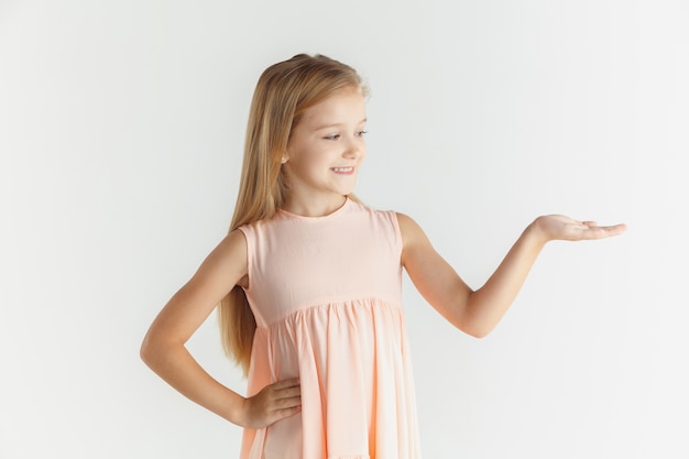 Stylish little smiling girl posing in dress isolated on white studio background. Caucasian blonde female model. Human emotions, facial expression, childhood. Showing on empty space bar.