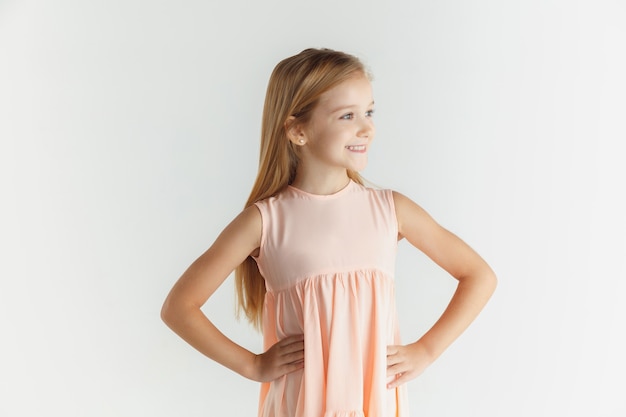 Stylish little smiling girl posing in dress isolated on white studio background. Caucasian blonde female model. Human emotions, facial expression, childhood. Smiling, holding hands on a belt.