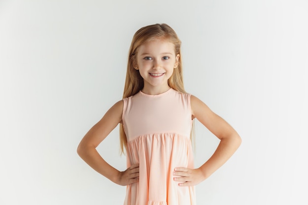 Stylish little smiling girl posing in dress isolated on white wall. Caucasian blonde female model. Human emotions, facial expression, childhood. Smiling, holding hands on a belt.