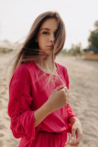 Stylish lovable female model in pink dress posing while outside photoshoot with flying har on deserted beach