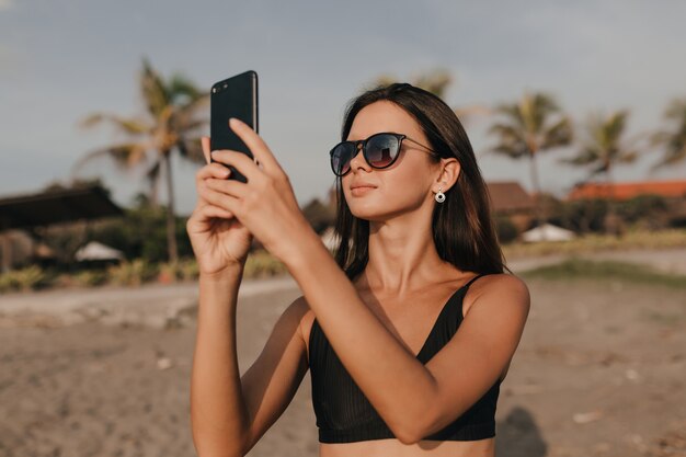 Stylish lovely woman with dark hair in sunglasses taking a picture on smartphone on the beach