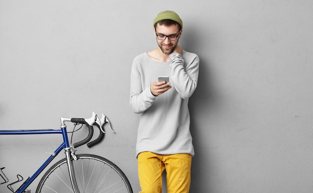 Стильный мужской велосипедист в зеленой шляпе и очках наслаждается онлайн-общением по мобильному телефону, его велосипед стоит рядом с ним