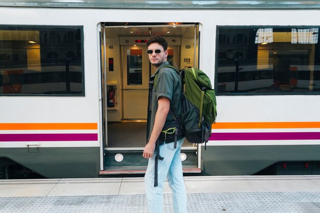 Stylish male tourist wearing eyeglasses and carrying backpack standing in front of railway train
