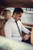 Free photo stylish male in a white shirt using smartphone in a luxury car.