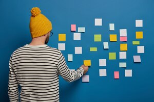 Stylish male youngster in fashionable outfit stands back to camera, turns to blue wall, sticks small colorful notes for putting down main ideas