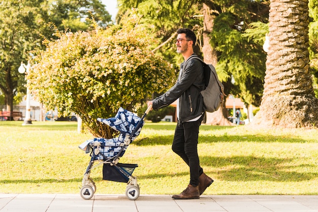 Zaino di trasporto alla moda dell'uomo che cammina con il passeggiatore di bambino nel parco