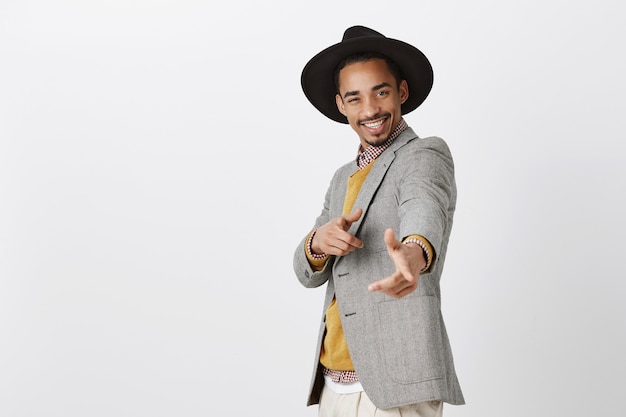 Stylish man charmed girls with his charisma. Indoor shot of good-looking stylish male in formal clothes and hat, pointing and smiling broadly, being in happy and good mood over gray wall