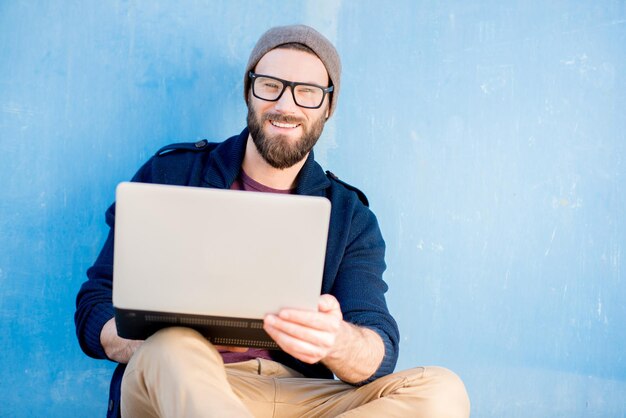Стильный мужчина, одетый небрежно в свитер и шляпу, работает с ноутбуком, сидя на фоне синей стены