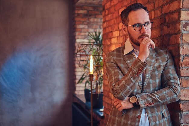 Stylish man in a flannel suit and glasses standing next to a against a brick wall