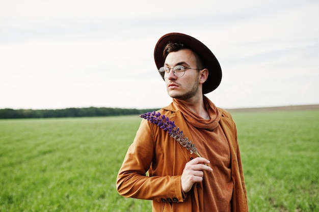 Stylish man in glasses brown jacket and hat posed on green field