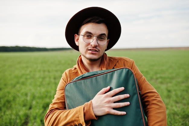 Stylish man in glasses brown jacket and hat with bag posed on green field